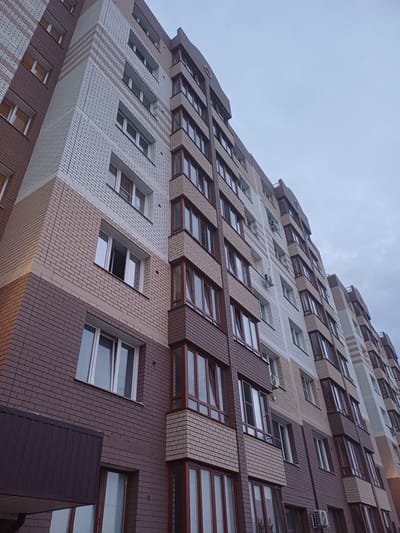 This is a low-angle, eye-level, full shot of a modern, multi-story building with a unique, stepped facade. The building is constructed with a mix of light tan, white, and dark brown brickwork, creating a visually dynamic pattern. Numerous windows are visible, many with dark brown frames, reflecting the overcast sky. Some windows are open, and a few air conditioning units are mounted on the exterior walls. The building's architecture features staggered sections and protruding elements, giving it an abstract, geometric appearance. The sky above is a pale, cloudy grey, suggesting an overcast day. No people are visible, and the scene appears to be an urban setting. The location is identified as Elitnyi, Russia.