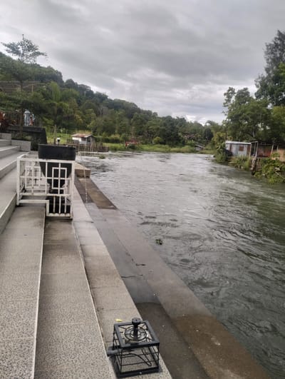 This image depicts a serene natural scene in Blang Bebangka, Indonesia, under a cloudy sky, suggesting an overcast or late afternoon setting. The primary focus is a wide, flowing river with ripples indicating a gentle current. On the left side of the image, a paved staircase leads down to the riverbank, featuring a decorative white metal railing and a large dark urn or planter from which a thin stream of water is cascading into the river. A small, black metal grate structure is also visible on the stairs.
The riverbank opposite the staircase is lined with dense green vegetation and trees, leading up to a gentle slope covered in foliage. Several small buildings, appearing to be simple houses or sheds with corrugated metal roofs, are situated along the riverbank on the right side. Some of these structures are elevated on stilts or foundations, a common adaptation in areas prone to flooding or near waterways. In the background, the landscape is dominated by lush, rolling hills covered in trees. The overall mood of the image is tranquil and natural, with a hint of rustic simplicity from the buildings. No people are clearly visible, and no text is present in the image.