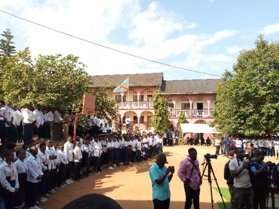 A large group of people, predominantly young adults, stand in a schoolyard in Beni, Democratic Republic of Congo.  The group is organized in rows, facing a white tent with a small group of people seated inside.  Several individuals wearing white shirts and black trousers are standing in front of the group of adults.  Two trees, one on each side of the frame, provide shade for the people gathered in the yard. There are several cameras set up on tripods with photographers taking pictures.  A basketball hoop is attached to a pole near the front of the group of adults, in front of the building.  The building in the background has a large, curved portico and a single Congo flag flying above the building.  The sky is clear and blue with a few scattered white clouds.  The ground is brown and dusty.  The scene is a formal gathering, likely a graduation ceremony, or a similar event.  The environment is hot and sunny with a dry, dusty atmosphere.  The location is in the city of Beni, Democratic Republic of Congo, at the coordinates (latitude: 0.492, longitude: 29.483).