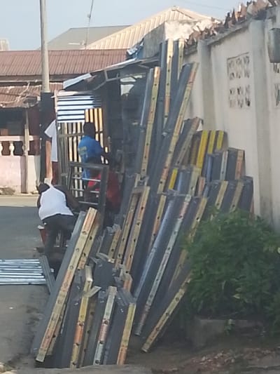 The image captures an outdoor scene in a daytime setting, likely a street or commercial area in Umuahia, Nigeria, showing activity related to construction materials.
In the foreground and midground, a substantial quantity of long, rectangular building materials is stacked upright against a wall on the right. These materials appear to be metal components, possibly roof trusses or support beams, characterized by alternating sections of dark grey/black and bright yellow/orange markings. A green potted plant or bush is visible at the bottom right, partially obscuring the materials.
Two men are present. In the lower-left quadrant, a man wearing a white tank top and dark trousers is bent over, seemingly engaged in work with a pile of silver corrugated metal sheets resting on the ground near him. Further into the midground, another man in a blue t-shirt stands behind a metal gate or grille structure, which appears to be part of a small shop or stall. A white t-shirt is hanging from this gate.
The background features several buildings with brown corrugated metal roofs, and a tall utility pole is visible on the left. A segment of a light-colored wall with decorative elements is seen on the far right. The overall scene suggests a busy working environment, likely involved in the sale or processing of building supplies under bright, clear weather conditions. No legible text is visible on any signs, clothing, or objects.