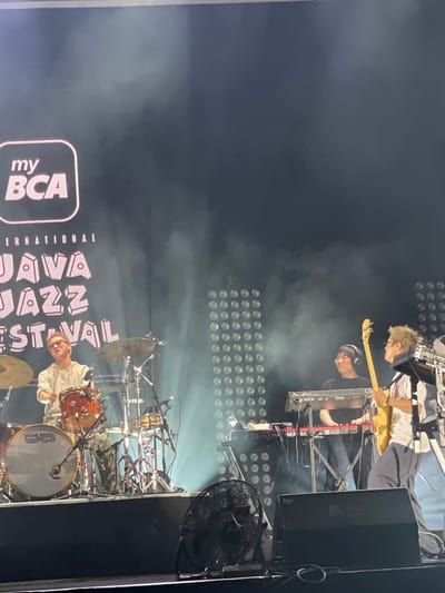 The image depicts a live musical performance at the Java Jazz Festival, presumably the BCA International Java Jazz Festival. The scene is likely set in Salembaran, Indonesia, based on the festival's name and origin. Three musicians are visible: a drummer, a keyboardist, and a guitarist/bassist. The drummer is seated behind a drum set, playing. The keyboardist is operating a red keyboard, and the guitarist is playing a light-colored guitar. Behind the performers, there is a wall of lights, and fog is present on stage. The "my BCA" logo is visible on the left side of the image, along with partial views of the "JAVA JAZZ FESTIVAL" banner. A black fan is visible stage right.