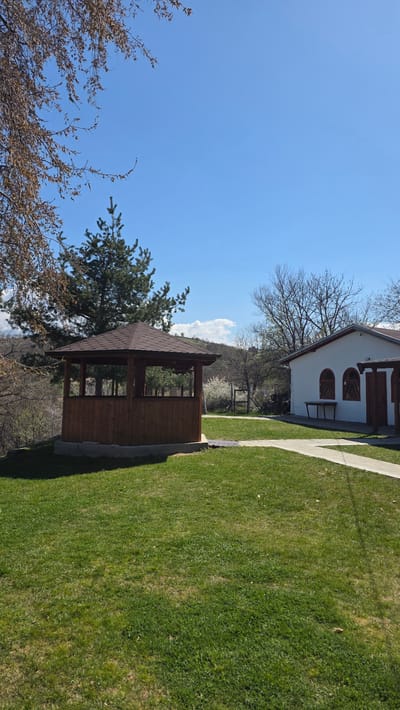 The image shows a view of a building and a gazebo in what appears to be a garden or park-like setting. The building, located on the right side of the image, is a white, single-story structure with dark red window frames. To the left of the building is a path made of stone, leading towards the back of the image. The gazebo is located on the left side of the image. It has a brown roof and is open on all sides, with a few trees surrounding it. The sky is blue. The Elenovo, Bulgaria location information, if correct, suggests this is the general location of the scene.
