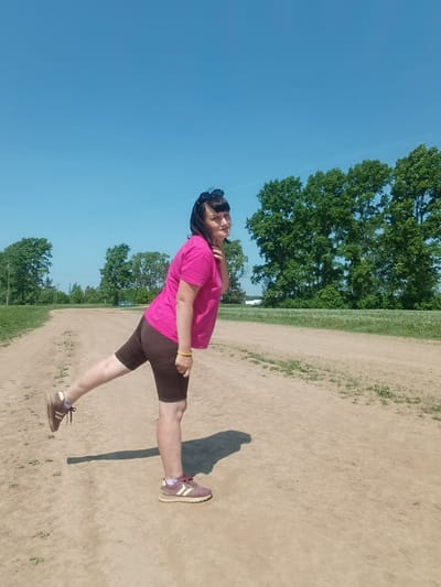 A woman stands on a dirt path, her left leg extended behind her, slightly bent at the knee. She wears a bright pink t-shirt, brown bike shorts, and maroon sneakers with light-colored stripes. Sunglasses are perched on top of her head. The sky above is clear and blue. Green trees line the horizon. The location is consistent with a rural or semi-rural setting, but without specific information, it is not possible to confirm or deny the presence in Foki, Russia.