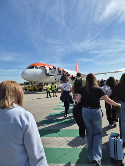 Passengers are boarding an EasyJet aircraft at an airport in Moreira, Portugal. The blue sky has a few white clouds. Some people are pulling their luggage. Airport personnel are assisting the passengers with boarding.