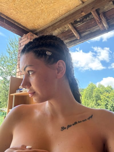 The image depicts a young woman with braided hair, wearing a nose ring and a tattoo on her upper chest that reads "She flies with her own wings." She is positioned outdoors, partially covering her breasts with her hand. The background reveals a wooden structure overhead, possibly a porch roof, with visible beams and insulation. Beyond this structure, lush green trees and a blue sky with white clouds are visible. The setting appears to be a rural or outdoor area, possibly a backyard or a campsite, consistent with a location in Belopoptsi, Bulgaria. The woman is looking off to the side, with her body angled towards the viewer. A shelf unit with bottles is visible to the left of the frame.