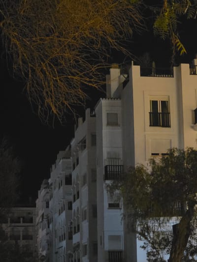 The image shows a nighttime view of apartment buildings in Ibiza, Spain. The architecture appears to be modern with white facades and dark balconies. The building on the right has several windows, some with balconies featuring black railings. The building on the left is less visible but also seems to be part of a residential complex with arched openings. Trees with bare branches and some foliage are in the foreground and partially obscure the buildings. The sky is dark, indicating it is nighttime. The lighting suggests artificial illumination from within the buildings or streetlights.
