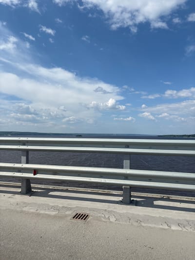 The image shows a view from a bridge overlooking a wide body of water. The water is dark and appears to be a river or lake. The sky is blue with scattered white clouds. In the foreground, there is a metal guardrail on the bridge, and a concrete surface with a drain grate. The shoreline on the other side of the water is visible in the distance, with trees and some buildings. The overall impression is a scenic, natural landscape. This scene could be located on one of the bridges crossing the Oka River in Orel, Russia, such as the Bridge of Lovers or the Central Bridge.