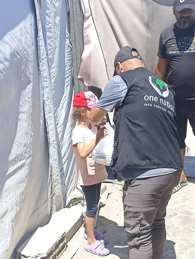 In the city of Gaza, a young girl wearing a pink t-shirt, dark leggings, and a red and pink cap receives an item from a person wearing a black vest with "one nation 100% donation policy" written on the back. The person in the vest is wearing a grey t-shirt underneath and dark pants. Another man wearing a black t-shirt with "NEVER STOP EXPLORING" and a black baseball cap stands in the background. They are in an outdoor area with a large, light-colored tarp or tent in the background and a stone surface below. The scene appears to be a distribution of goods or aid.