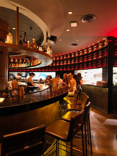 The image shows the interior of a dimly lit, modern bar. A circular bar dominates the foreground, with bottles of alcohol displayed on shelves behind it. Several people are seated at the bar, including a woman in a black tank top who appears to be the bartender, as she is seen working behind the bar. A group of women are seated at the bar, engaged in conversation. The bar is well-stocked with various spirits and other beverages, and there are bottles of wine neatly arranged on shelves in the background. The overall ambiance is sophisticated and upscale, with warm lighting and a warm color palette. The scene is set in Be'er Sheva, Israel. There is no visible text in the image.