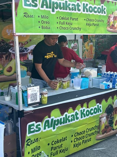 The image shows a street vendor in Makassar, Indonesia, selling "Es Alpukat Kocok," a blended avocado drink. The vendor, wearing a black t-shirt, is preparing drinks behind a counter laden with ingredients and serving cups. The stall features a vibrant green banner with the product name and a list of flavors, including Milo, Oreo, Boba, Coklat Parut, Full Keju, and Choco Crunchy Keju Parut. Various containers of ingredients, such as shaved ice, milk cartons, and cups, are visible on the counter. A QR code is also displayed for payment. In the background, a person in a red shirt is visible, along with other stalls and greenery, suggesting an outdoor market or street food setting. The overall scene depicts a typical urban food stall operation.