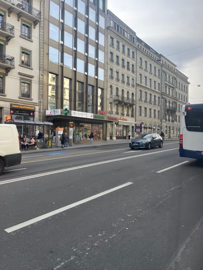 This media file captures a street scene in Geneva, Switzerland, under an overcast sky during daytime. The image is oriented vertically but depicts a horizontal street view.
On the right, an asphalt road features white lane markings, with a partial view of the rear of a white and blue bus, bearing the name "GITARD." Further down the road, a dark-colored car is visible, possibly parked or moving slowly.
The left side of the scene is dominated by urban architecture. A tall, multi-story building displays classical European design with numerous windows, decorative cornices, and ornate balconies on its upper floors. At street level, various shops occupy the ground floor. Visible signage includes "PHARMACIE" (French for Pharmacy) and "Parapharmacie," indicating a pharmacy. Another prominent sign reads "TABAC CHANTEPOULET" (Tabac referring to a tobacco shop) and "Souvenirs," distinctly featuring the Swiss cross emblem, which helps confirm the Geneva location, as "Chantepoulet" is a street in the city. Adjacent to this, a more modern building with a facade of large, reflective glass windows is also visible.
Several pedestrians are present on the sidewalk. Some individuals are seen near the shop entrances, while others are walking or waiting near what appears to be a bus stop area. One person is positioned in the street near the dark car. The overall setting depicts a typical busy urban environment with a blend of historical and contemporary buildings.