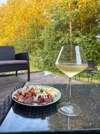 In an outdoor setting with trees and foliage in the background, a table is set for a meal. On a plate are slices of meat and cubes of cheese. To the right of the plate is a glass of white wine. In the foreground, a dark wicker chair is visible. The scene appears to be in Zamosh'e (Zaklinskoe S/P), Russia, given the context.