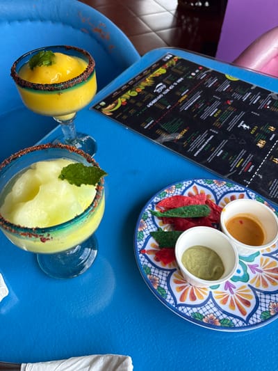 The image shows a blue table with two margaritas and a plate of chips and dip. The margaritas are both in tall, stemmed glasses. The one on the left is green and slushy. The one on the right is yellow and more liquid. The plate has chips in red and green, and two dipping sauces. One sauce is a light green and the other is orange. There is a menu on the table to the right of the drinks, and a white napkin to the left. The table is located at a restaurant in Pozos, Costa Rica, at the coordinates 9.951, -84.194.