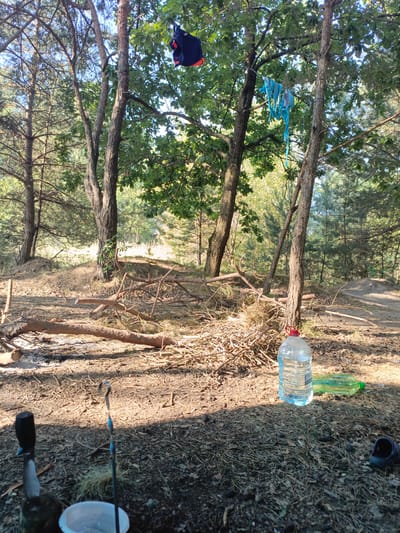 In the city of Мухавецкі сельскі Савет, Belarus, the image depicts a campsite scene in a wooded area. A large plastic bottle of water stands in the foreground, accompanied by a green plastic bottle and a dark blue sandal. In the middle ground, a pile of dry branches and logs suggests preparation for a fire, with remnants of ash visible. Overhead, a dark blue garment and a blue bikini are suspended from a wire strung between trees, indicating laundry is drying. The environment is characterized by trees, dry ground cover, and dappled sunlight filtering through the foliage.