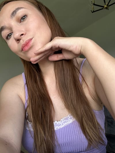This is a close-up, vertical, selfie-style photo of a person's face and upper body. The person has long, reddish-brown hair that falls over their shoulder and chest. They are wearing a ribbed, lavender-colored tank top with white lace trim. Their skin appears fair and smooth, and they have green eyes. Their left hand is raised and gently touches their cheek and mouth area. Their lips are a soft pink. The background is a plain, muted green wall. A portion of a black and gold chandelier-style light fixture is visible in the upper left corner. The lighting suggests it could be daytime indoors. The overall mood of the image is calm and introspective. There is no visible text in the image.
