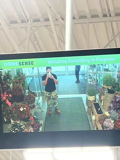 A surveillance monitor displays a security camera feed inside a HomeSense store located in the Town of Cortlandt, United States of America. The feed shows a man with light skin wearing a black beanie, black shirt, and camouflage pants, taking a picture with his phone. The scene also captures other individuals further in the background and various decorative plants and flower arrangements near the foreground. The monitor indicates that recording is in progress.