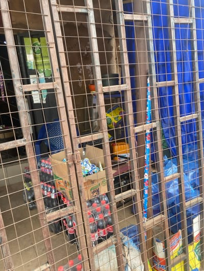 The image depicts a closed storefront in Okura, Nigeria, viewed through a heavily rusted, brown metal security gate. The gate, formed by a grid of vertical and horizontal bars, is closed and occupies the foreground. Behind the gate, the interior of a retail space is visible. Stacks of plastic bottles, primarily Coca-Cola, are arranged on the floor and in cardboard boxes, including one branded "Peak." A blue plastic chair is positioned on the left. A partially visible green sign with "gloo" branding and "VIDEO AVAILABLE HERE" text is affixed to the left interior wall. Further inside, a dark container rests on a shelf, and a yellow jerry can is visible. A long, slender object with a blue background and red floral patterns stands upright on the right. The background on the right side is dominated by a large blue tarp or fabric, behind which bags, one labeled "SUPREME," are discernible. No individuals are present in the scene.