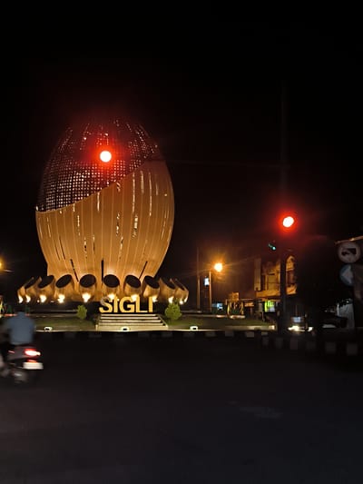 An eye-level, medium shot captures a night scene at a roundabout in the city of Lampeudeu Baroh, Indonesia. The focal point is a large, egg-shaped monument made of golden, slatted metal with a checkered pattern on top. The monument is illuminated from within and by lights at its base. The word "SIGLI" is displayed in large, golden, three-dimensional letters on a stepped platform in front of the monument. To the right of the monument, a traffic light displays a red signal. In the foreground, a dark asphalt road runs across the bottom of the frame. On the left side of the road, a person on a motorcycle is blurred due to motion, heading towards the right. The background is dark, with some buildings and streetlights visible. The overall impression is one of a well-lit urban landmark at night.