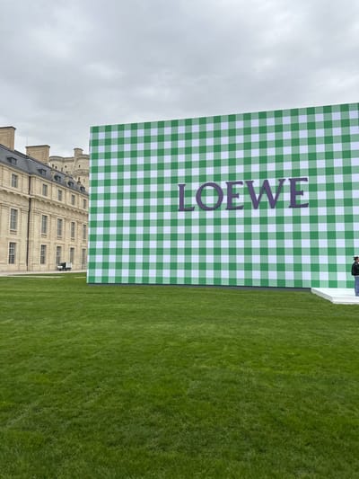 The image captures an outdoor scene at the Château de Vincennes, France, under an overcast sky. A large, temporary structure, approximately two stories tall, stands prominently in the midground to the right, covered in a vivid green and white gingham pattern. The word "LOEWE" is displayed in large purple capital letters across its surface. A single individual is partially visible on the far right, standing on a low, white platform at the base of the gingham structure, facing leftward. The foreground consists of a meticulously maintained expanse of lush green grass. To the left, a historical multi-story building, characteristic of the Château de Vincennes complex with its light-colored stone facade and multiple windows, runs horizontally. Beyond this building, in the distant background, a fortified stone structure, likely a tower or keep of the Château, is visible. A small white utility vehicle is parked on a paved area to the left, between the lawn and the historic building. The overall scene suggests a set-up for a public event or presentation.
