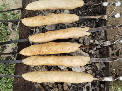 Six uncooked sausage-like items are skewered on metal rods and are arranged horizontally above glowing charcoal. The items appear to be made of ground meat or a similar mixture, and they have a pale, off-white color with some slight browning in places. The skewers are resting on a metal grill over a bed of charcoal. The background consists of dirt and some sparse green vegetation. The scene appears to be an outdoor cooking setting, possibly in a yard or park. There are no people visible in the image. The location is specified as Зелёное, Russia.