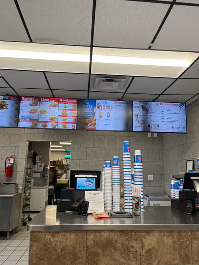 This is an interior shot of a Dairy Queen restaurant in Johnson City, Tennessee, United States of America. The image focuses on the ordering counter area. Above the counter are digital menu boards displaying "Blizzard Menu", "Treats", "Shakes and Malts", "Drinks", "More Eats", "Baskets", and "Sides". The menus feature prices for various Dairy Queen items. Several stacks of Dairy Queen branded cups are on the counter. A cash register, a customer-facing screen displaying the "Blizzard" logo, and a payment terminal sit on the counter. A poster indicating a "Free Dilly Bar" incentive is visible. Behind the counter is a partial view of the kitchen area, including shelving with supplies and a fire extinguisher. The general color palette is neutral with tile walls and a textured ceiling with recessed lighting.