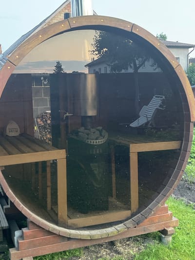 The image shows a wooden barrel sauna located outdoors in the city of Jelgava, Latvia. The sauna has a large, transparent glass front. Inside the sauna, a wood-fired stove with stones is visible, along with wooden benches. To the left, a pile of firewood is stacked against a brick wall. A white lounge chair is positioned inside on the right side. The sauna is set on a wooden base, surrounded by grass and garden vegetation. The background shows residential buildings and trees. The sky is visible through the glass front and appears to be overcast. There are no people visible in the image.