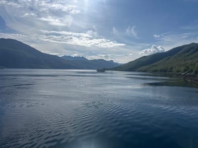 The image shows a wide, calm body of water, likely a fjord, with steep, tree-covered mountains on either side. The sky is blue with scattered white clouds, and the sun is shining, creating reflections on the water. In the distance, a small ferry boat is visible. The overall impression is one of a serene and beautiful natural landscape. The image was taken from the water, looking towards the shore. The location is Inndyr, Norway.