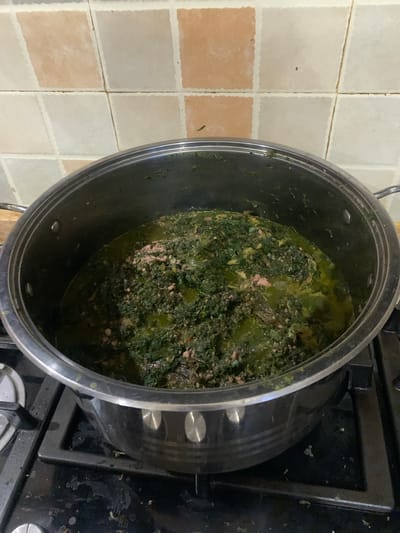 A high-angle, medium shot captures a large, stainless steel pot filled with a dark green, leafy stew containing small pieces of meat, situated on a black gas stove. The stew appears to be a traditional Nigerian dish, possibly Ewedu soup or Ogbono soup, commonly prepared in homes across Matogbun, Nigeria. The background consists of a tiled wall with square tiles in shades of cream and peachy-orange, a common kitchen feature in Nigerian households. The stovetop is slightly soiled, indicating recent use. The overall scene depicts a domestic setting, likely a kitchen, where a meal is being prepared.
