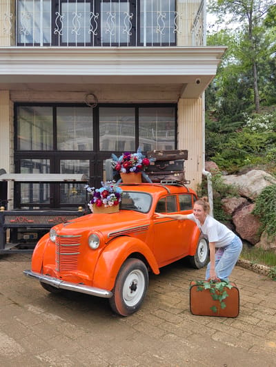 The image depicts a young woman posing next to a vintage orange car, likely for a photograph, in the Ukrainian city of Luchistoe. The car is an older model, possibly a Moskvich, decorated with flower-filled baskets on the roof and topped with a stack of vintage suitcases. The woman is standing with one hand on the car and another holding a brown suitcase adorned with ivy. The location appears to be a paved area in front of a two-story building with a decorative facade. There's a wrought-iron structure or bench to the left of the car. The background includes trees and rocks, suggesting a semi-rural or park-like setting. A security camera is visible on the building.