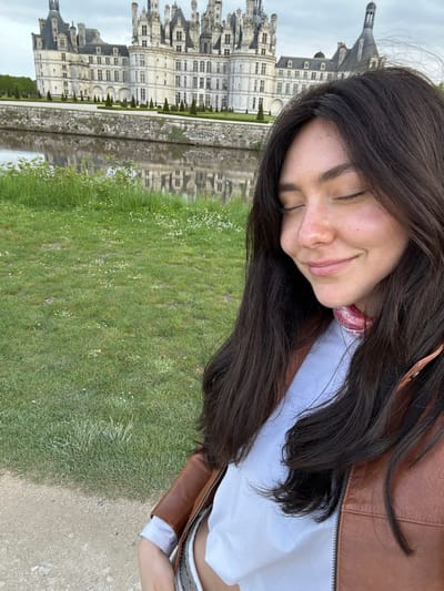 The photo shows a woman with long, dark hair, wearing a brown leather jacket and a white T-shirt with a pink and red bandana around her neck. She is standing in front of the Chateau de Chambord, a grand Renaissance castle in the Loire Valley region of France. The coordinates 47.617, 1.515 indicate the location of the castle. The woman is standing on a path next to a moat. She is looking down at the ground and smiling. Her eyes are closed. The castle is visible in the background. The day is overcast and the water is still. The ground is covered in grass and flowers. The scene is serene and peaceful.