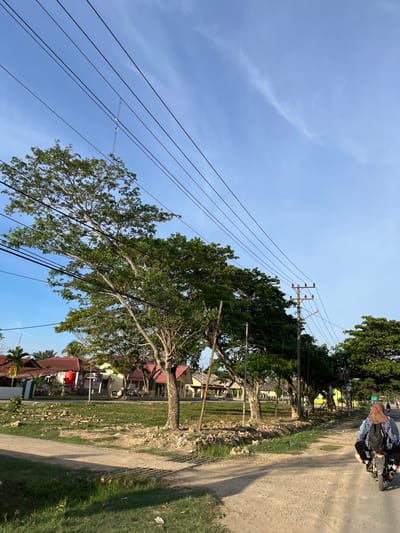 The image shows a dirt road lined with trees and power lines, with houses visible in the background. A person is riding a motorcycle down the road, wearing a headscarf and backpack. The sky is blue with some wispy clouds. The scene appears to be a rural or suburban area in Alurcucur, Indonesia. The lighting suggests it is late afternoon or early morning.