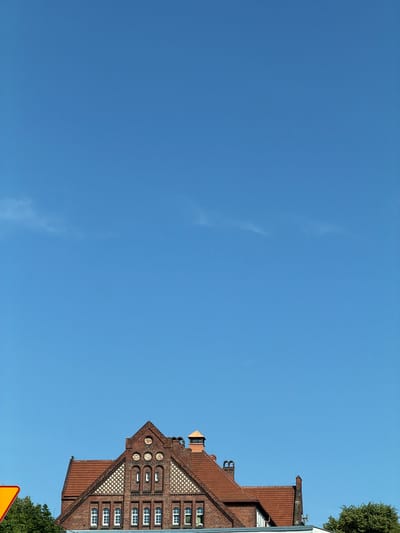 The image shows the upper facade of a brick building with a red tile roof, set against a clear blue sky. The building features several rows of windows with white frames. The architectural style appears to be historical, with decorative brickwork and arched window detailing. A chimney is visible on the roof. To the left of the building, a portion of a yellow triangular sign is visible, and green trees are present. The image is taken from a low angle, looking upwards. The location is Gdańsk, Poland, specifically in the area of the former Railway Station Administration Building, now the Polish Baltic Philharmonic.