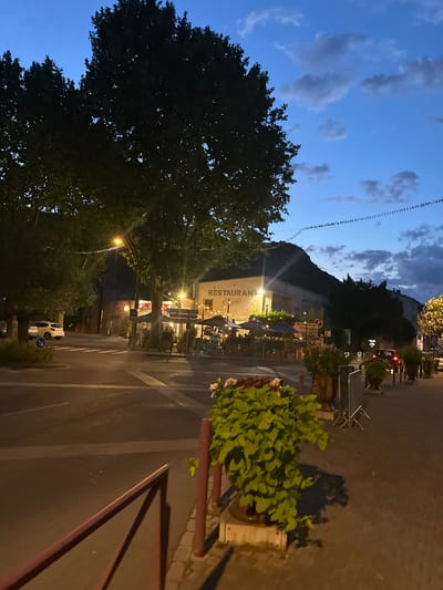 The image depicts a street scene at dusk in Anduze, France. The sky is a deep blue with scattered clouds, suggesting the transition from day to night. A string of decorative lights hangs across the street, adding a festive atmosphere.

On the right side of the street, there are potted plants and outdoor seating for a restaurant. The restaurant's facade is visible, illuminated by warm lights. A sign on the building reads "RESTAURANT" and "PIZZA", indicating the type of establishment. Further down the street, another sign with "RESTAURANT" is also visible.

On the left side of the street, a large tree with dark foliage dominates the foreground. Beyond the tree, a hillside rises, and a building is nestled into its slope. The street itself appears to be wet, possibly from recent rain or to keep the dust down. A white car is parked on the side of the road.

The scene is quiet and peaceful, with a sense of a warm evening in a European town. The lighting suggests it is late evening, and the atmosphere is relaxed, perhaps with diners enjoying their meals or people strolling through the town. There are no people clearly visible in the image. The overall impression is one of a charming and inviting place to be during the evening.