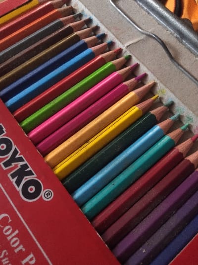 Approximately 25 hexagonal colored pencils, exhibiting sharpened tips and minor use, are arranged diagonally in two rows within a red TOYKO® brand cardboard box. The visible pencil colors include shades of yellow, orange, pink, red, green, blue, brown, black, and purple. The pencils are held in place by a light brown cardboard insert. The red box flap prominently displays the black "TOYKO® Color P" logo, with additional text partially visible as "Sur". Adjacent to the box, on a light brown, textured surface, a silver-colored metallic U-shaped object is partially visible, alongside a blurred orange object. The scene is located in Жарсуат, Kazakhstan.