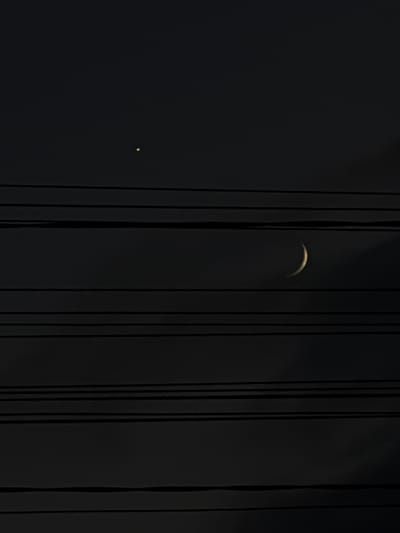 The image shows a dark sky with several horizontal lines that appear to be power lines. In the upper left portion of the sky, a small, bright yellow point of light is visible, likely a planet or star. In the middle right of the sky, a crescent moon with a faint glow is also visible. The overall scene is one of a night sky with celestial bodies and man-made infrastructure. No people or specific landmarks are visible. The location in Dongaron, Russia cannot be determined from the image alone.