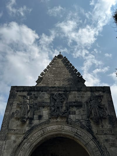 The image shows the exterior facade of the San Andrés Apóstol church in San Andrés Calpan, Mexico, from a low-angle perspective, looking upwards towards a blue sky with white clouds. The church features intricate stone carvings. Prominently displayed are two angelic figures with wings, one on the left and one on the right, flanking a central relief carving of what appears to be a saint or warrior figure. Above these figures, text is inscribed in stone, with "S. MIGUEL" visible on the left and "S. MIGUEL ARCANGEL" on the right. Below the central relief, another inscription reads "S. MIGUEL". The facade is adorned with decorative stonework, including an arched entryway at the bottom. The architecture suggests a historical and religious context.