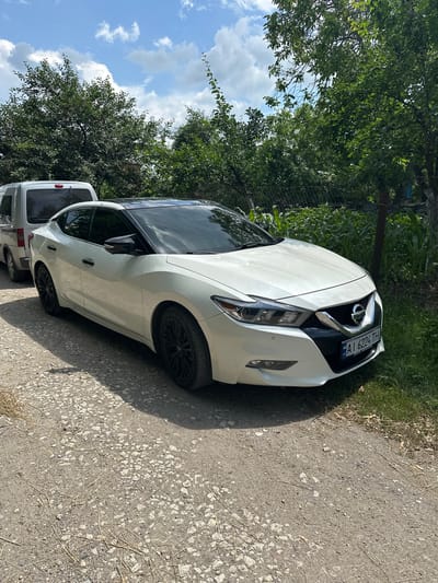This media file shows a white Nissan sedan parked on a gravelly road. The car is positioned at an angle, with its front end facing towards the right of the frame. The car has a black roof and black rims on its wheels. Behind the Nissan, a white van is partially visible.

The setting appears to be outdoors, with lush green trees and vegetation lining the left side of the road. The sky is blue with scattered white clouds, suggesting it is daytime and the weather is pleasant. The road surface is rough, composed of gravel and small stones, with dry grass and twigs scattered across it.

A license plate is visible on the front of the Nissan, displaying the text "AL 6294 TH" in a blue and yellow format. This license plate is likely from Ukraine, given the location context of Horodyshche-Pustovarivske. There is no indication of any people or specific activities taking place in the scene. The overall impression is one of a parked vehicle in a rural or semi-rural environment on a sunny day.