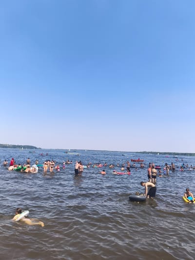 The media file depicts a crowded summer day at a lake, likely in Nieporęt, Poland. A large number of people are enjoying recreational activities in the water under a clear, bright blue sky, suggesting fair weather and midday. The scene is bustling with activity, with individuals swimming, floating on inflatable devices, and some appearing to be sunbathing or socializing in the water.
Subjects include a diverse group of people of various ages, many wearing swimwear. Inflatable objects such as tubes, rings, and floats are prominent, aiding relaxation and play. Boats and kayaks are also visible in the distance, indicating broader water sports are taking place. The setting is a wide expanse of water, with a tree-lined shoreline visible in the background, further confirming the lakeside location.
The overall mood appears to be one of leisure and enjoyment, with people of all ages participating in water-based activities. The bright sunlight and clear sky contribute to a cheerful and lively atmosphere. There is no visible text in the image.