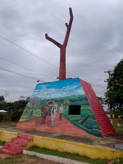 This image features a distinctive monument located in Concepción, Paraguay, captured on an overcast day. The central structure consists of a large, reddish, pyramid-shaped base from which a tall, stylized tree-like sculpture, also reddish and textured, extends upwards into the sky.

The base of the monument is covered by a detailed mural depicting a rural scene. On the left side of the mural, two oxen, one brown and one white with black markings, are shown yoked together. Alongside them walks a man wearing a straw hat and light clothing, holding a stick. The painted background features lush green hills and distant mountains beneath a bright blue sky with white clouds, suggesting a typical pastoral landscape. A pink flowering tree and a smaller tree with yellow foliage are also part of the mural's scenery. Integrated into the painting is a dark, rectangular opening, possibly a window or a recess in the monument itself, and below it, a dark cylindrical object resembling a cannon barrel. Faint text near the cannon includes a signature and date, appearing to be "ARTURO [...] 2022". The unpainted portions of the monument's base are textured and red.

The monument is situated in an outdoor public space. In the foreground, on the left, a set of red concrete steps with a yellow painted trim leads up to the structure. Overhead, multiple black power lines are visible against the grey, cloudy sky. To the right, a utility pole supporting a street light and green leafy trees are present, contributing to the public park or town square setting. The atmosphere is calm, with the overcast weather suggesting a mild day.
