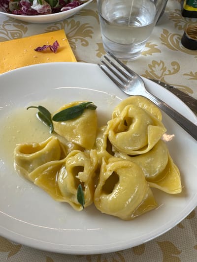 The image displays a close-up, elevated view of a dining table setting in Anzola dell'Emilia, Italy. In the foreground, a white, oval ceramic plate holds a portion of approximately eight to ten light-yellow tortelloni or tortellini, glistening with a light-colored, possibly butter-based, sauce. Several small, fresh green sage leaves are placed on the pasta.
To the right of the pasta plate, a silver-colored dining fork with four tines rests diagonally across its rim, with a portion of a matching knife handle visible underneath. Above the fork, a clear glass tumbler filled with water, exhibiting condensation, is positioned. To the right of the water glass, the dark-colored cap of a bottle, possibly containing olive oil, is partially visible.
To the left of the main plate, a folded yellow paper napkin is visible, upon which lie a few small white particles and a single reddish-purple radicchio leaf fragment. In the upper-left background, a partial view of a white bowl containing a salad of mixed greens, predominantly red radicchio and green lettuce, is present. The entire arrangement is situated on a light-colored tablecloth featuring a repeating pattern of gold or light brown stylized floral/scroll motifs. The scene depicts a prepared meal in an indoor dining environment; no individuals are present.