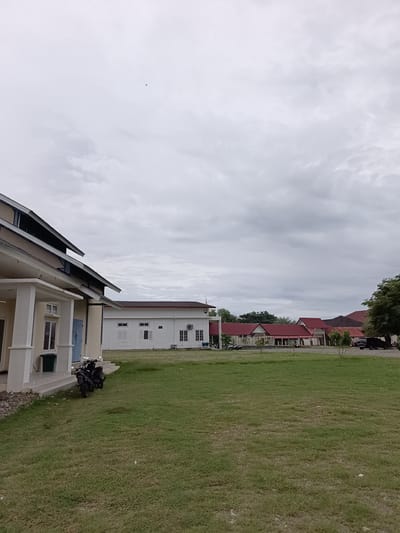A medium-shot, eye-level, outdoor, wide-angle photograph captures a building complex under a cloudy sky in Mesjid Reuntoh, Indonesia. The foreground features a neatly mown, green lawn, with a few scattered small white objects. Two dark-colored motorcycles are parked on a paved area to the left, near a light-colored, multi-story building with a tan facade, white columns, and a dark sloped roof. To the right of this building, a white, single-story structure with a dark roof has several windows with shutters and an air conditioning unit visible. Further back, a row of buildings with red, sloped roofs and white walls stretches across the frame. In the distance, a large green tree stands on the far right, and a dark-colored vehicle is parked near the red-roofed buildings. The sky is overcast with dense gray clouds. There are no people visible in the image.