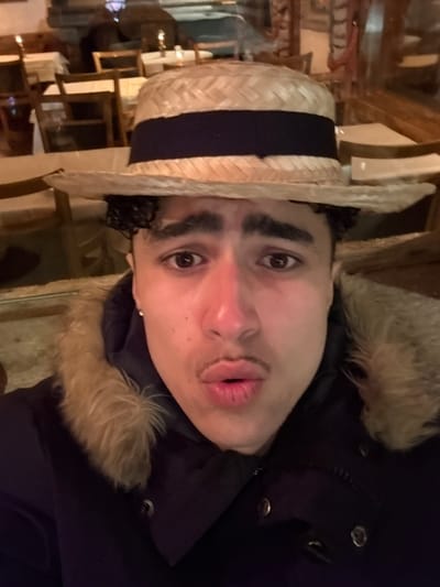 An individual is depicted in a close-up, chest-up frame in the city of La Clusaz, France. The person wears a straw boater hat with a dark band, a dark winter coat featuring a prominent light brown fur-lined hood, and a dark hoodie beneath. Visible features include dark curly hair, a short mustache above pursed lips forming an 'O' shape, wide-open eyes, and a silver stud earring in the left ear. The background consists of reflections showing an indoor environment, specifically what appears to be a restaurant or cafe interior with wooden chairs, tables covered with white tablecloths, and small illuminated objects, possibly candles or lamps.