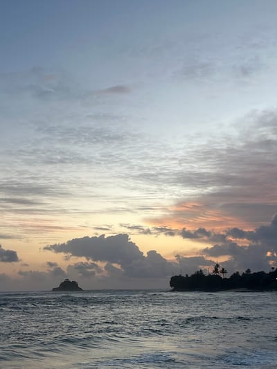 The image depicts a scenic sunset over the ocean in Midigama, Sri Lanka. The sky is filled with a dramatic display of clouds, ranging from soft, diffused white to darker, more textured gray and purple formations. Hints of warm, golden and pink hues from the setting sun are visible through the clouds, creating a serene and beautiful atmosphere.

The scene features a silhouette of a tropical coastline on the right side of the image. Several palm trees are visible against the colorful sky, indicating a beach or coastal location. The ocean occupies the right half of the frame, with its surface textured by waves, reflecting the muted light of the sunset. The water appears calm, with gentle waves rolling towards the shore.

The time of day is clearly sunset, characterized by the soft, warm light and the changing colors in the sky. The weather appears to be partly cloudy, adding visual interest to the sky. The location is identified as Midigama, Sri Lanka, a known surf destination, which is consistent with the presence of ocean waves and palm trees.

There is no visible text in the image. The overall mood conveyed by the image is peaceful, tranquil, and awe-inspiring, capturing the natural beauty of a tropical sunset.