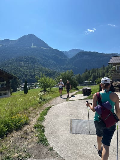 In Schönau am Königssee, Germany, on a clear, sunny day, two individuals are walking away from the camera on a paved path. The person in the foreground, wearing a white baseball cap, turquoise tank top, and dark shorts, is using trekking poles. A red backpack with a purple towel draped over it is slung over their shoulder. Behind them, two other individuals, both appearing to be male, are walking on the same path. They are wearing athletic clothing and backpacks. The path winds through a grassy area with wildflowers and trees. In the background, a lush green mountain covered in dense forest rises towards a bright blue sky. Several traditional Bavarian-style houses with wooden facades are visible nestled among the trees and meadows. The scene depicts a scenic hiking or walking trail in a mountainous region.