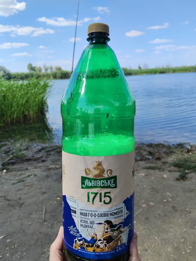 This media file features a close-up of a large green plastic bottle of Lvivske 1715 beer, held by a hand. The bottle has a label with text and graphics. The label indicates the beer is "Lvivske 1715" and features the text "НАШІ Г-О-О-ОЛОВНІ МОМЕНТИ" (Our Goal Moments) and "УСПІХ, ЩО НАДИХАЄ" (Success that Inspires). There is also a date "26 ЧЕРВНЯ 2006" (June 26, 2006) visible. The beer is described as "світле пастеризоване" (light pasteurized) with 4.5% alcohol by volume.
The background suggests an outdoor setting near a body of water, possibly a lake or river. Tall green reeds are visible on the left bank, and the water itself is calm with ripples. The ground in the foreground is sandy or muddy. A fishing rod can be seen in the background, extending upwards against a blue sky dotted with white clouds. The lighting indicates it is daytime, likely late morning or early afternoon. The overall mood is relaxed and casual, suggesting a leisurely outdoor activity like fishing or picnicking. There is no indication of the location being Lima, Peru; the text on the bottle is in Ukrainian.