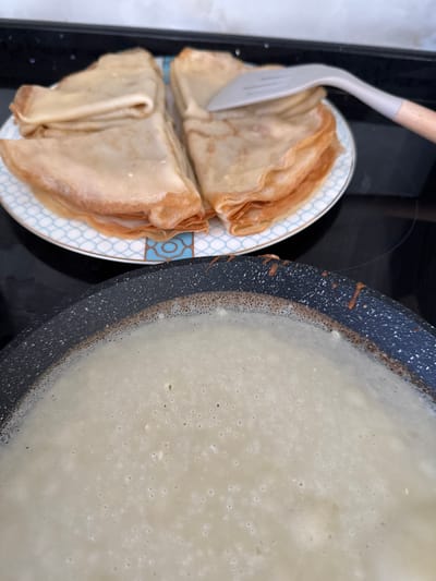 In Uyutnoye, Ukraine, on a kitchen stovetop, a pile of golden-brown crepes is stacked on a white and blue patterned plate. A grey silicone spatula with a wooden handle is partially covering the crepes on the right side. In the foreground, a black non-stick frying pan is filled with a light-colored batter, likely for more crepes, with small bubbles forming on the surface. The background shows a dark stovetop surface.