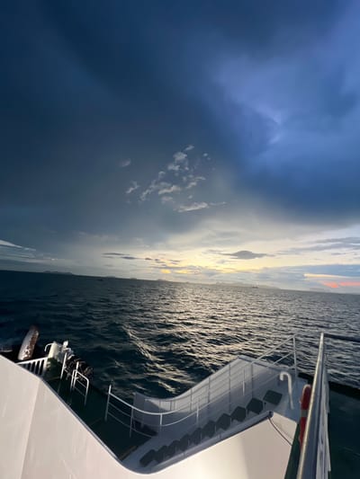 An exterior, eye-level, medium shot captures a ferry deck at sea during dusk. The foreground is dominated by the white structure of the ferry, with railings and stairs visible. The sea, with moderate waves, stretches out to the horizon, where distant islands are silhouetted against a dramatic sky. The sky is a mix of dark, stormy clouds in the upper left and lighter, warmer tones towards the horizon, with hints of orange and yellow as the sun sets. There are no people visible in the frame. The overall impression is one of a journey at sea as daylight fades.