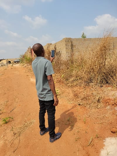 The image, captured in Nwofia, Nigeria, displays an adult male standing on reddish-brown unpaved ground. The subject is viewed from the back, slightly to the right, and is wearing a grey short-sleeved polo shirt, black jeans, and dark blue loafers. His right arm is raised, holding a dark-colored smartphone vertically, oriented upwards and slightly to the right. In the mid-ground, a partially constructed wall composed of light-colored concrete blocks extends horizontally. Dense, dry, yellowish-brown vegetation is present against and around this wall. Further in the background to the left, a pile of building blocks and a light-colored vehicle are partially visible. The foreground and mid-ground consist of dry, reddish-brown earth with sparse green sprouts. The sky is light blue with scattered white cumulus clouds. A pronounced shadow of the man is cast on the ground to his right.