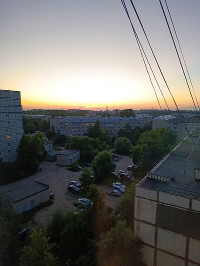 This is a high-angle, eye-level shot of a residential area in Alexandrov, Russia, during what appears to be sunset or sunrise. The sky is a gradient of pale yellow and orange near the horizon, transitioning to a muted blue-gray overhead. Silhouetted against the sky are power lines, running diagonally across the frame from the top right.
The scene is dominated by several multi-story apartment buildings, typical of Soviet-era architecture. The closest apartment building on the left is tall and gray with visible window frames. Another apartment building, further into the background, has a blue tint and lit windows, suggesting occupancy. Other, lower buildings, possibly garages or small industrial structures, are scattered among the trees.
A parking lot is visible with several cars parked, including a red sedan, a dark hatchback, and several other vehicles in various colors. Lush green trees and foliage are interspersed throughout the scene, indicating a time of year with active vegetation, likely summer. The overall impression is of a quiet, everyday urban environment bathed in the soft light of dawn or dusk. There is no visible text in the image.