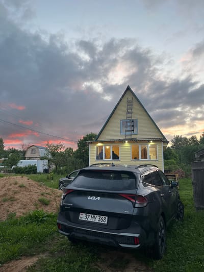 In Клобушнево, Russia, a dark gray Kia Sportage is parked on a grassy area in front of a two-story yellow house. The house has several windows illuminated from within and a ladder leaning against its peaked roof. Behind the house, more trees and a greenhouse are visible. A large pile of sand is to the left of the house, and another car is parked in the background. The sky is dramatic with dark clouds streaked with vibrant pink and orange hues from a sunset or sunrise. The scene appears to be a rural residential area.